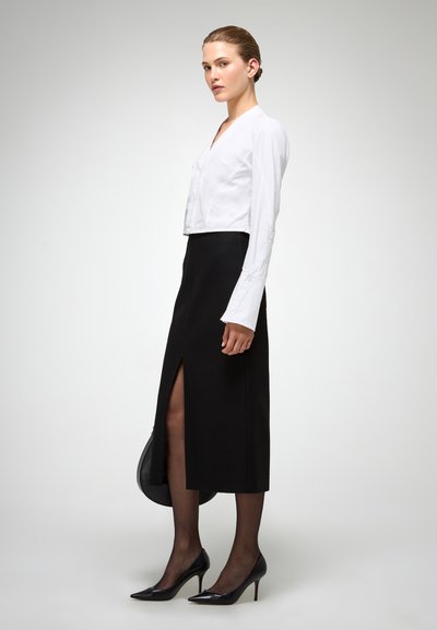 Cropped blouse bianca abbinata a una gonna midi nera con uno spacco alto fino alla coscia. La modella indossa collant neri trasparenti e décolleté neri a punta.