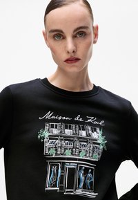 Svart hettegenser med en butikkillustrasjon med grønne detaljer, vinduer og figurer. Tekst "Maison de Kol" i hvitt over designet.