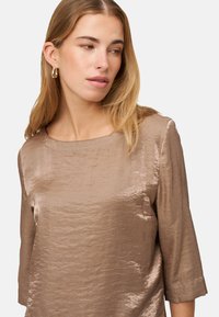 Bronzefarbene Bluse mit glatter, glänzender Textur, rundem Ausschnitt und Dreiviertelärmeln. Minimalistisches Design ohne sichtbare Muster.