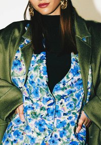 Robe à motif floral aux nuances de bleu et de vert, portée avec un col roulé noir sous un manteau vert olive. Caractéristiques : col et boutons.