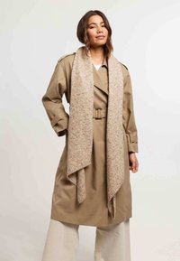 Femme portant un trench-coat beige ceinturé, un pantalon large crème et une longue écharpe en tricot beige, posant avec une main sur la hanche.