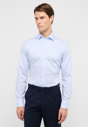Camicia da uomo a righe azzurro chiaro in cotone, con colletto button-down, bottoni dorati e maniche lunghe. Indossata con pantaloni blu navy.