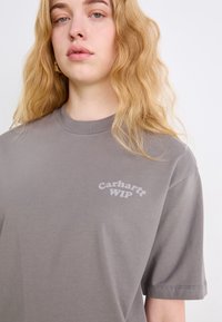 Maglietta grigia a maniche corte in cotone, con un logo sottile in testo grigio chiaro sul petto e collo a giro.