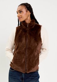Femme avec des cheveux tressés portant un gilet en fausse fourrure marron à fermeture éclair, un haut à manches longues blanc et un jean bleu, les mains dans les poches du gilet, regardant à gauche.
