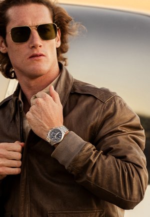 Veste bombardier en cuir marron avec des poignets côtelés, chemise grise et montre en argent avec un cadran noir et un bracelet en métal. Lunettes de soleil teintées en vert.