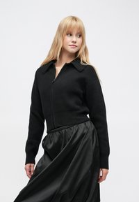 Pulover croșetat negru, scurt, cu fermoar frontal, cu guler ascuțit și manșete striate, asortat cu o fustă din satin negru, fluidă.