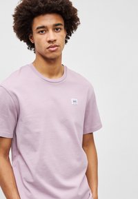 Ανοιχτό μωβ βαμβακερό T-shirt με στρογγυλή λαιμόκοψη και κοντά μανίκια. Διαθέτει μια μικρή μπλε και λευκή ετικέτα στο στήθος. Λείο υλικό.