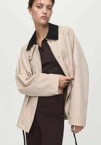 Femme portant une veste beige avec un col noir sur un haut côtelé marron foncé et un pantalon marron foncé, ajustant le zip de la veste.