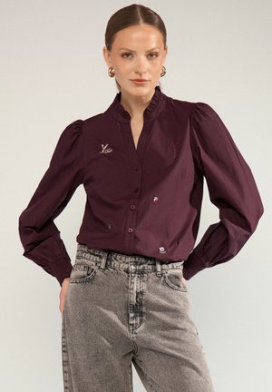 Vrouw die een donkerpaarse blouse met knopen en pofmouwen draagt met kleine geborduurde motieven, gecombineerd met hooggetailleerde vervaagde grijze jeans.