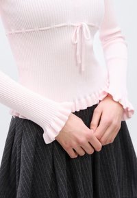Haut côtelé rose clair à manches longues avec poignets à volants, doté d'un détail noué à la taille. Associé à une jupe rayée foncée.