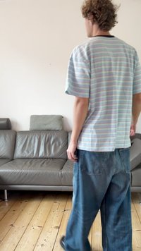 Srajca s kratkimi rokavi z modro, roza in belo črtami v pastelnih odtenkih, kombinirana z ohlapnimi modrimi kavbojkami. V ozadju siva usnjena sedežna garnitura.