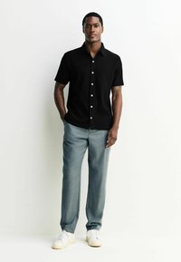 Sort kortærmet skjorte med et tekstureret mønster og hvide knapper, sammen med lysegrå bukser og hvide sneakers.