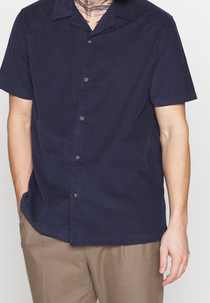 Homme portant une chemise bleu marine à manches courtes boutonnée et un pantalon beige, une main dans la poche, sur un fond uni.