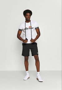 Bijela majica kratkih rukava s crnim "ellesse" logotipom i prugastim rukavima, u kombinaciji s crnim šortsevima i bijelim Nike tenisicama.
