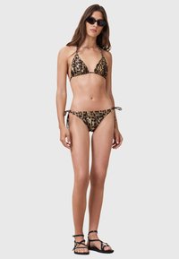 Set bikini con stampa animale in tonalità di marrone e nero, caratterizzato da un top triangolare e un fondo con lacci laterali. Il modello indossa occhiali da sole neri e sandali.