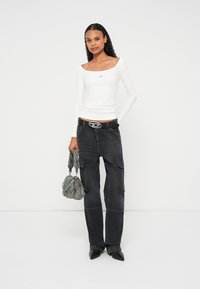 Weißes Langarmshirt mit rundem Ausschnitt, kombiniert mit dunklen Cargo-Hosen und einer grauen, strukturierten Handtasche. Schwarze spitze Stiefel vervollständigen das Outfit.