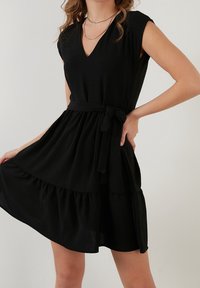 Svart kort klänning i strukturerad tyg, med v-ringning, korta ärmar, ett knytbälte i midjan och en lagerdesign på kjolen.