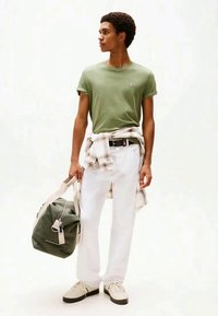 Camiseta verde de algodão, calças brancas e sapatilhas bege. Segurando uma bolsa de desporto verde com um puxador de fecho azul e uma camisa aos quadrados atada à cintura.