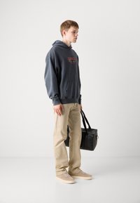Sudadera gris con bolsillo frontal, pantalones beige y zapatillas beige. Sostiene una bolsa negra con doble asa. Tejido suave y corte relajado.