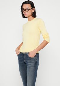 Ανοιχτό κίτρινο πλεκτό πουλόβερ με μακριά μανίκια, στρογγυλή λαιμόκοψη και ριμπ μανσέτες, συνδυασμένο με σκούρο μπλε εφαρμοστά τζιν.