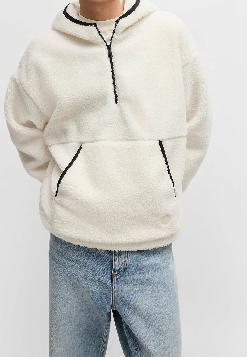 Personne portant un sweat à capuche en polaire blanche avec une fermeture éclair noire et un bord de poche, associée à un jean denim bleu clair, debout devant un fond uni.