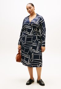 Φόρεμα ναυτικού μπλε με μοτίβο λευκών πινελιών, σχεδιασμός τύπου κρουαζέ, μακριά μανίκια, σφηκοφωλιά στη μέση με κυκλική ζώνη, συνδυασμένο με καφέ τσάντα και μαύρα παπούτσια.