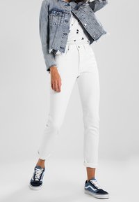 Veste en denim bleu clair avec un ourlet effiloché, jeans blancs ajustés, et baskets bleues avec des accents blancs. Chemise blanche à motifs en dessous.