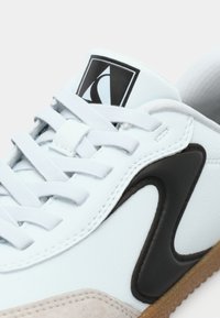 Baskets blanches avec un détail noir en forme de vague, bout en daim beige clair, lacets blancs et logo noir et blanc sur la languette.