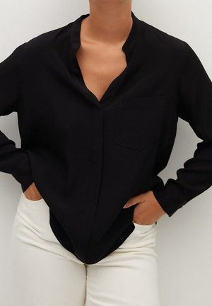 Persona che indossa una camicetta nera a maniche lunghe senza colletto e pantaloni bianchi, con le mani nelle tasche dei pantaloni.