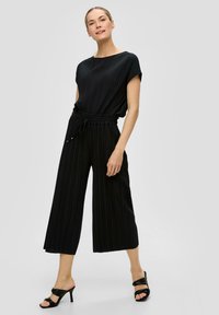 Zwarte geplooide jumpsuit met korte mouwen, elastische tailleband en wijde, cropped pijpen. Gecombineerd met zwarte hoge muiltjes.