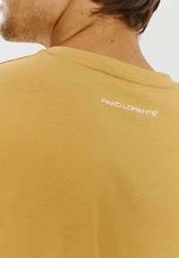 Żółta bawełniana koszulka z okrągłym dekoltem, pokazująca gładką fakturę i małe białe logo marki z tyłu, blisko dekoltu.