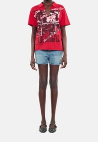 Polo shirt rouge à manches courtes avec impression graphique et texte, associé à un short en denim. Modèle portant des ballerines noires à clous.