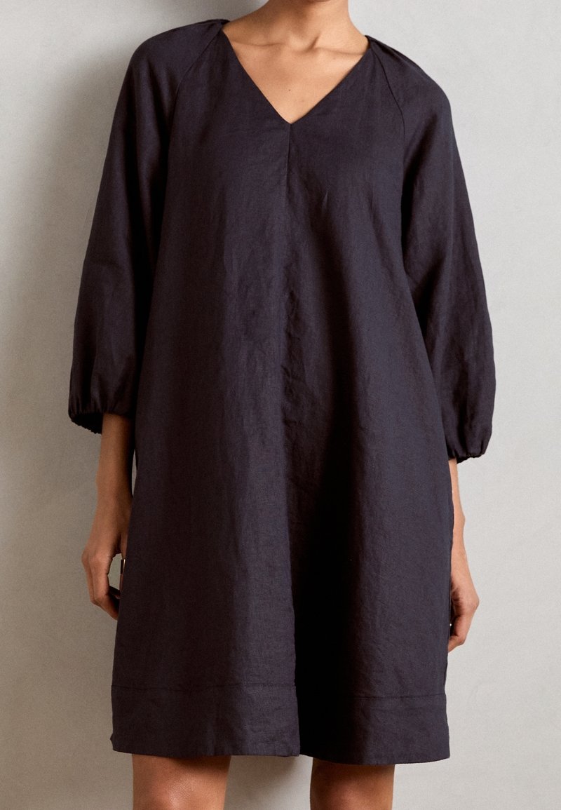 Femme portant une robe noire ample, longueur genou, avec des manches trois-quarts et un col en V, sur fond uni.