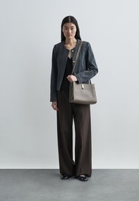 Pelēka strukturēta žakete ar zelta pogām, melns tops, platas brūnas bikses un pelēks pleca soma ar metāla aizdari, uz neitrālas fona.