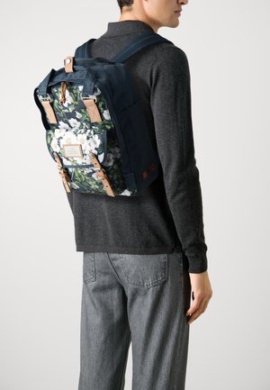 Une personne portant un sac à dos marine avec un motif floral blanc et des sangles en cuir beige, un pull gris foncé et un jean gris clair, de profil.