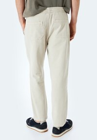 Pantalones de algodón beiges con un corte relajado, cinturilla elástica, dos bolsillos traseros y dobladillos ajustados, combinados con zapatillas oscuras.