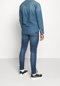 Džínová košile na knoflíky ve střední modré barvě, v kombinaci s tmavě modrými skinny džíny a černými teniskami s bílými podrážkami, pohled zezadu.