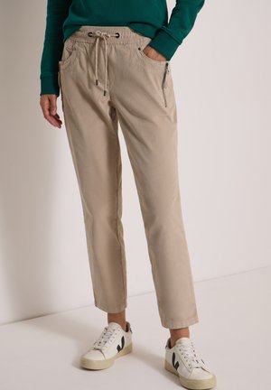 Femme portant un pantalon beige à cordon avec poche zippée et des baskets blanches, debout devant un fond uni.