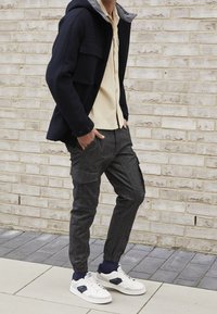Marineblauwe capuchonjack met grijze voering, beige knoopshirt en donkergrijze cargobroek met taps toelopende pijpen. Witte sneakers met marineblauwe accenten.