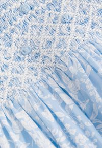 Robe bleu clair avec corsage froncé orné de motifs floraux blancs, tissu texturé et jupe plissée. Comprend des boutons décoratifs.