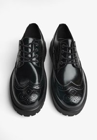 Scarpe in pelle nera con finitura lucida, punta rotonda e design wingtip. Presentano dettagli perforati, accenti cuciti e una suola spessa e testurizzata.
