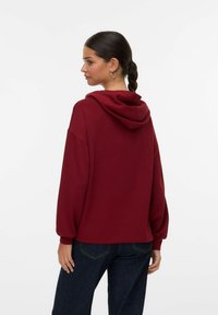 Sudadera con capucha color burdeos de corte holgado, con hombros caídos y puños acanalados, combinada con jeans de mezclilla oscuros. Textura suave.