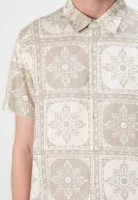 Kortermet skjorte laget av lett stoff, med et beige og elfenbensfarget blomsterdesign, knappestolpe og nedfellbar krage.
