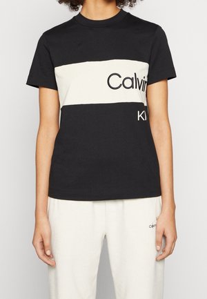 Kobieta ubrana w czarno-białą koszulkę w blokowe kolory z częściowym logo "Calvin Klein" oraz białe spodnie z małym detalem logo.