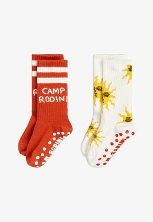 Meias de algodão vermelhas com riscas brancas e texto "CAMP RODINI"; meias brancas com padrão de sol amarelo e bolinhas vermelhas na sola.