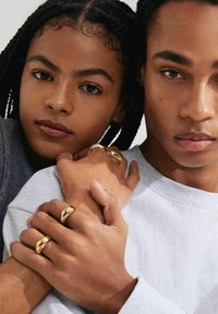 Goldene Ringe mit glatten, geschwungenen Formen schmücken die Hände. Eine Hand trägt einen grauen Pullover, während die andere nackt ist. Der Hintergrund ist hell und neutral.