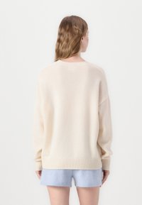 Lys beige oversized genser laget av mykt stoff, med ribbestrikket hem og mansjetter. Bært over lyseblå shorts, sett fra baksiden.