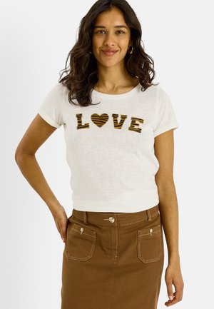 Femme aux longs cheveux foncés portant un t-shirt blanc avec «LOVE» en lettres rayées et une jupe marron avec des poches devant, souriant à la caméra.