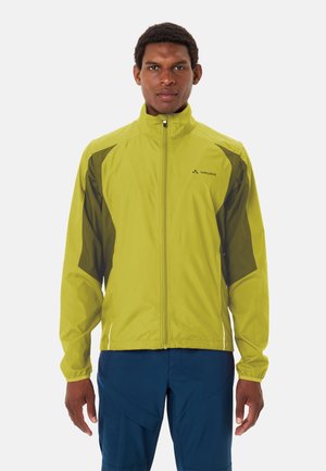 Man die naar voren kijkt en een limegroene en donkergroene jas met rits draagt, gecombineerd met een blauwe broek, tegen een witte achtergrond.