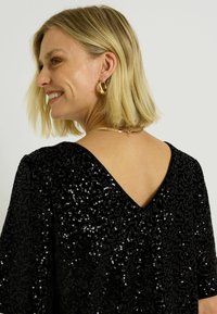 Haut noir à sequins avec un décolleté en V, manches courtes et surface texturée, mettant en avant des sequins scintillants qui créent de la profondeur visuelle.